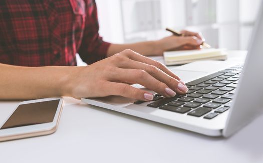 Gut gepflegte Hände arbeiten an einem Laptop und machen Notizen in einem offenen Notizbuchvvvvvvvvvvvvvvvv